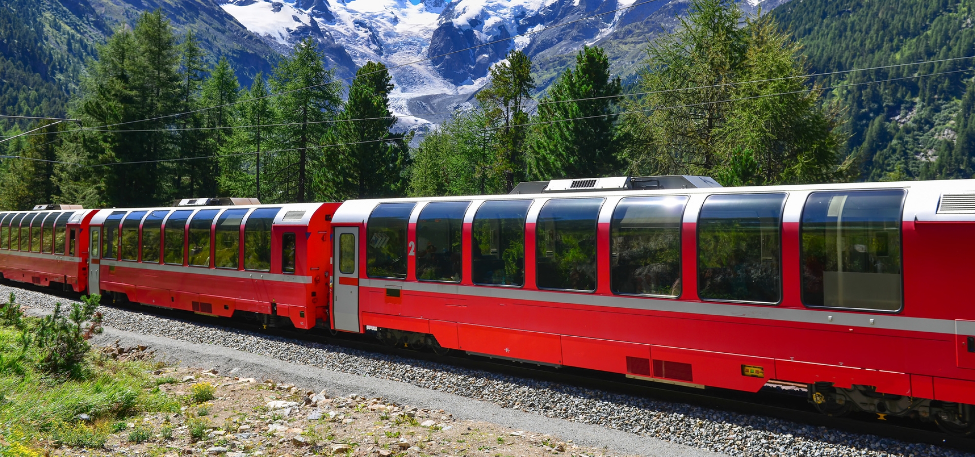 A train travels through Alpine scenery with bright blue sky